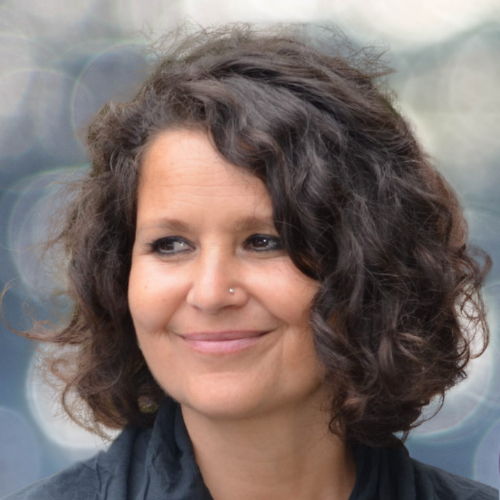 Smiling woman with short curly dark hair and a nose stud, wearing a dark jacket outdoors.